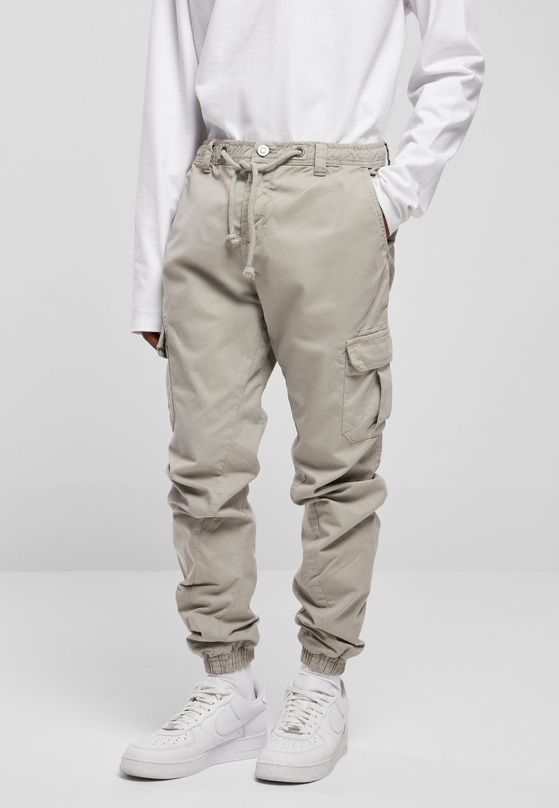 Beige Cargo-Hosen mit einem elastischen Bund, verstellbarem Kordelzug, elastischen Knöchelbündchen und mehreren Seitentaschen. Getragen mit weißen Sneakern.