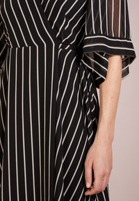 Mustavalkoinen raidallinen mekko, jossa on kietaisumallinen suunnittelu, läpinäkyvät hihat, sidottava vyötärö ja pehmeä materiaali. Sisältää pystysuorat raidat visuaalisen pituuden lisäämiseksi.