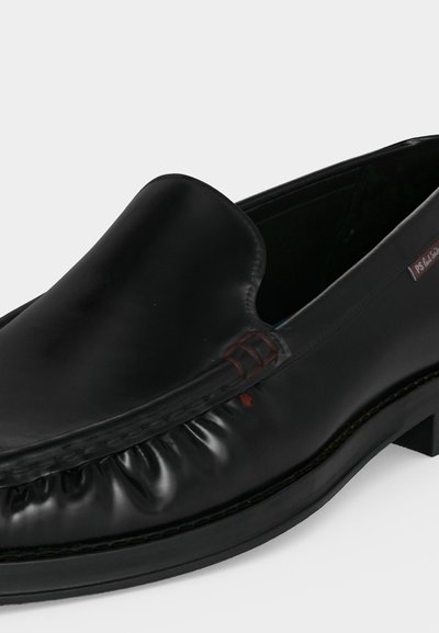 Mocassin noir en cuir, à enfiler, avec bout arrondi, coutures visibles et petite étiquette rouge sur le côté, présenté sur un fond uni.