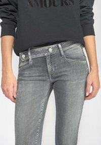 Osoba v bledě šedých džínách s vysokým pasem, které mají knoflík a zip, v kombinaci s černou mikinou s textem "AMOURS".