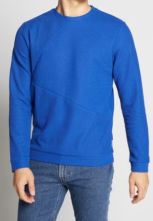 Mand iført en blå, langærmet tekstureret pullover-striktrøje kombineret med blå denimjeans, stående foran en ensfarvet baggrund.