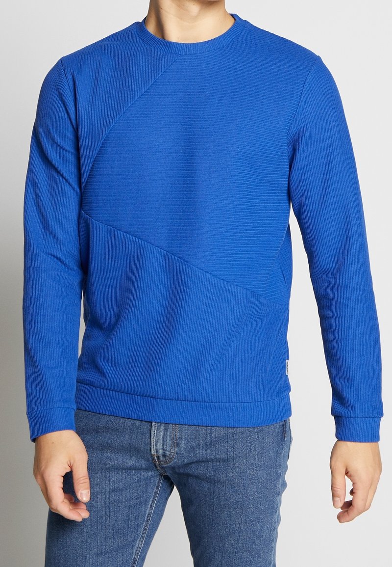 Homme portant un pull à manches longues bleu texturé associé à un jean en denim bleu, debout devant un fond uni.