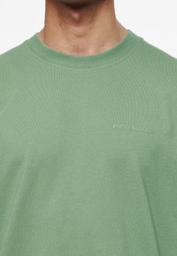 Zelené bavlněné tričko s kulatým výstřihem a krátkými rukávy. Na hrudi má jemné logo "PEGADOR" ve stejné barvě.