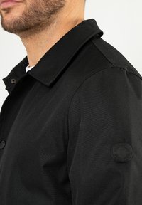 Czarna kurtka zapinana na guziki z wyraźnym kołnierzem, teksturowanym materiałem i okrągłą naszywką z logo na lewym rękawie. Zbliżenie na ramię.