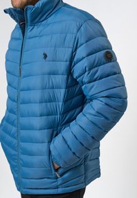 Homme portant une veste matelassée bleue zippée, mains dans les poches, avec un logo noir brodé sur la poitrine et un patch circulaire sur la manche.