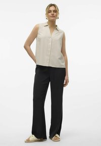 Chemise sans manches beige avec un devant à boutons, associée à un pantalon à jambes larges noir et des sandales de couleur claire, offrant une coupe décontractée.
