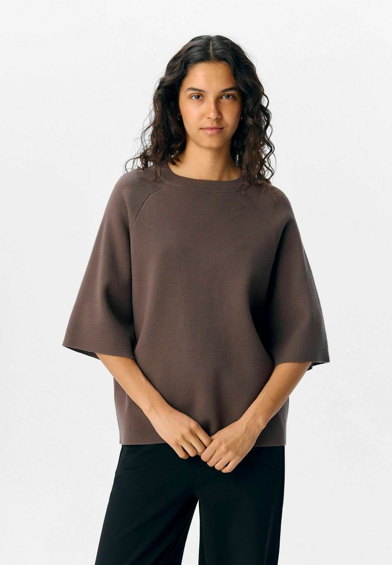 Pull en mailles brun avec un col rond, des manches courtes et une coupe décontractée, associé à un pantalon noir.