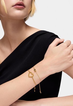 Pulsera de oro con forma de corazón, con cuentas redondas y una cadena. Lleva puesta en una muñeca desnuda sobre una blusa negra. Textura suave.