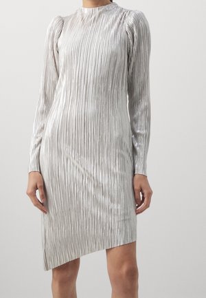Femme portant une robe plissée argentée à manches longues, col haut et ourlet asymétrique sur un fond clair uni.