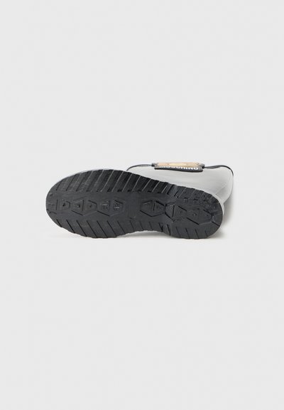 Semelle en caoutchouc noir d'une botte présentant un motif texturé, avec un détail de boucle beige et un branding visible sur la sangle.