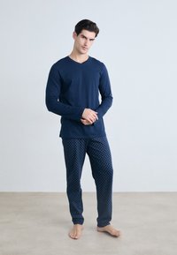 Hombre con camiseta de manga larga azul marino y pantalones azul marino con estampado, de pie descalzo frente a un fondo gris claro liso.