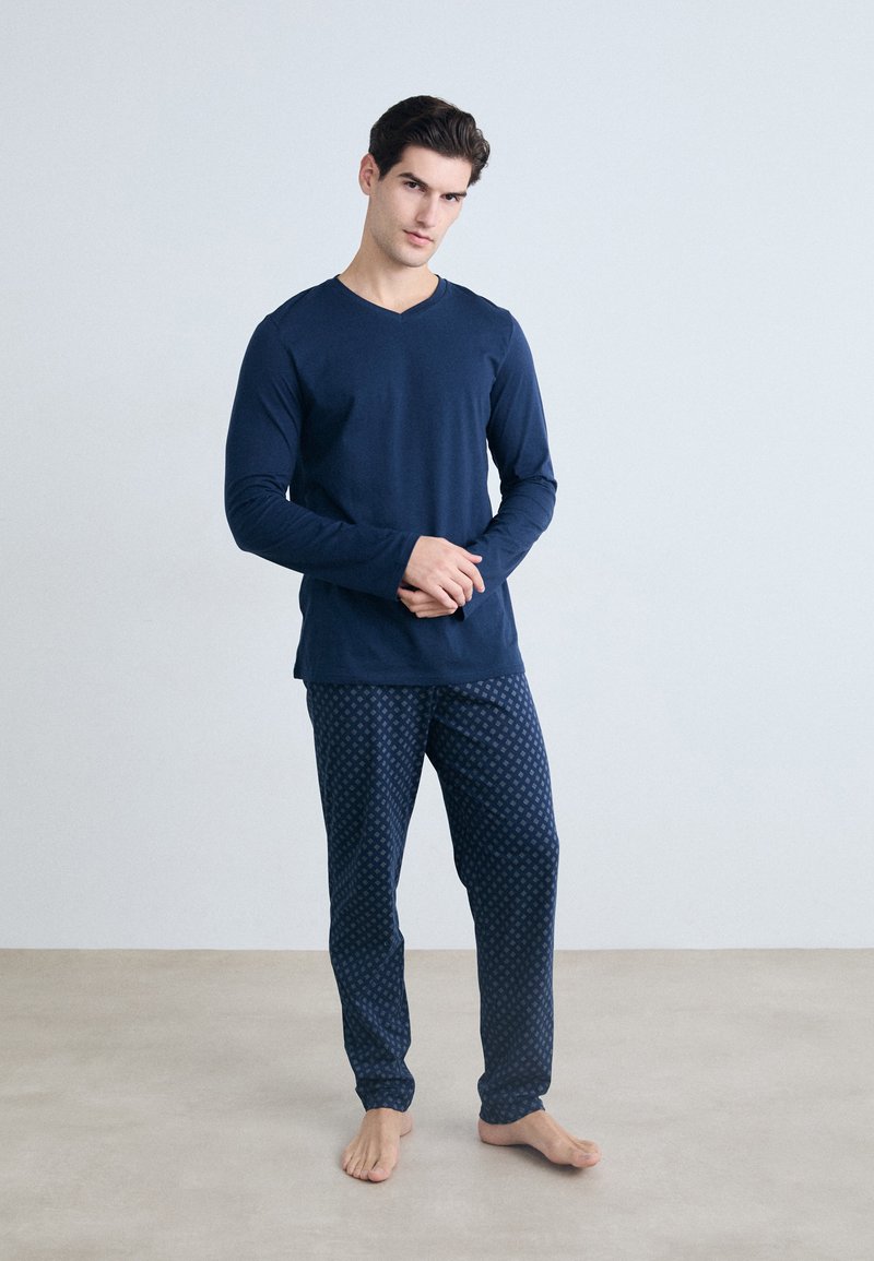 Hombre con camiseta de manga larga azul marino y pantalones azul marino con estampado, de pie descalzo frente a un fondo gris claro liso.