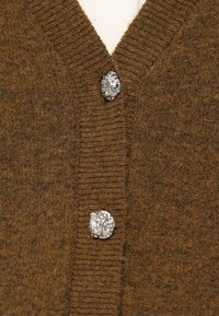 Cardigã castanho de lã com dois grandes botões prateados irregulares fixados sobre uma peça de vestuário branca por baixo.