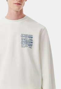 Sudadera blanca con un pequeño logo rectangular azul que presenta el texto repetitivo "LACOSTE" en el lado izquierdo del pecho. Diseño de mangas largas y cuello redondo.