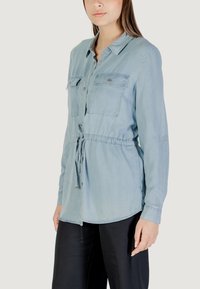 Camisa de manga larga azul claro con dos bolsillos en el pecho, cierre de botones en la parte delantera, cintura ajustable con cordón y dobladillo curvado. Textura de tela suave.