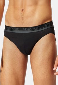 Männlicher Oberkörper mit schwarzen Unterwäsche, die einen grauen Bund mit der Aufschrift "uncover" trägt, vor einem schlichten hellen Hintergrund.