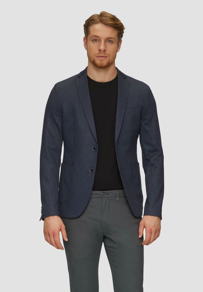 Blazer a cuadros en tonos navy, confeccionado con tejido texturizado, con un corte ajustado, solapas de muesca y dos bolsillos delanteros. Combinado con una camisa negra y pantalones grises.