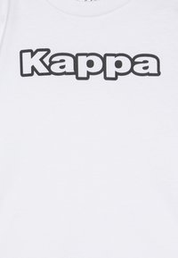 Bela bomba majica iz bombaža z imenom blagovne znamke "Kappa" v velikih, krepkih, črnih črkah na sprednji strani. Klasična zasnova s krožno ovratnikom.