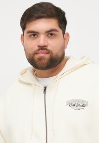 Kremfarget zip-hoodie laget av mykt stoff, med logo-trykk på venstre bryst og snorer på hetten. Enkel design, uten mønstre.