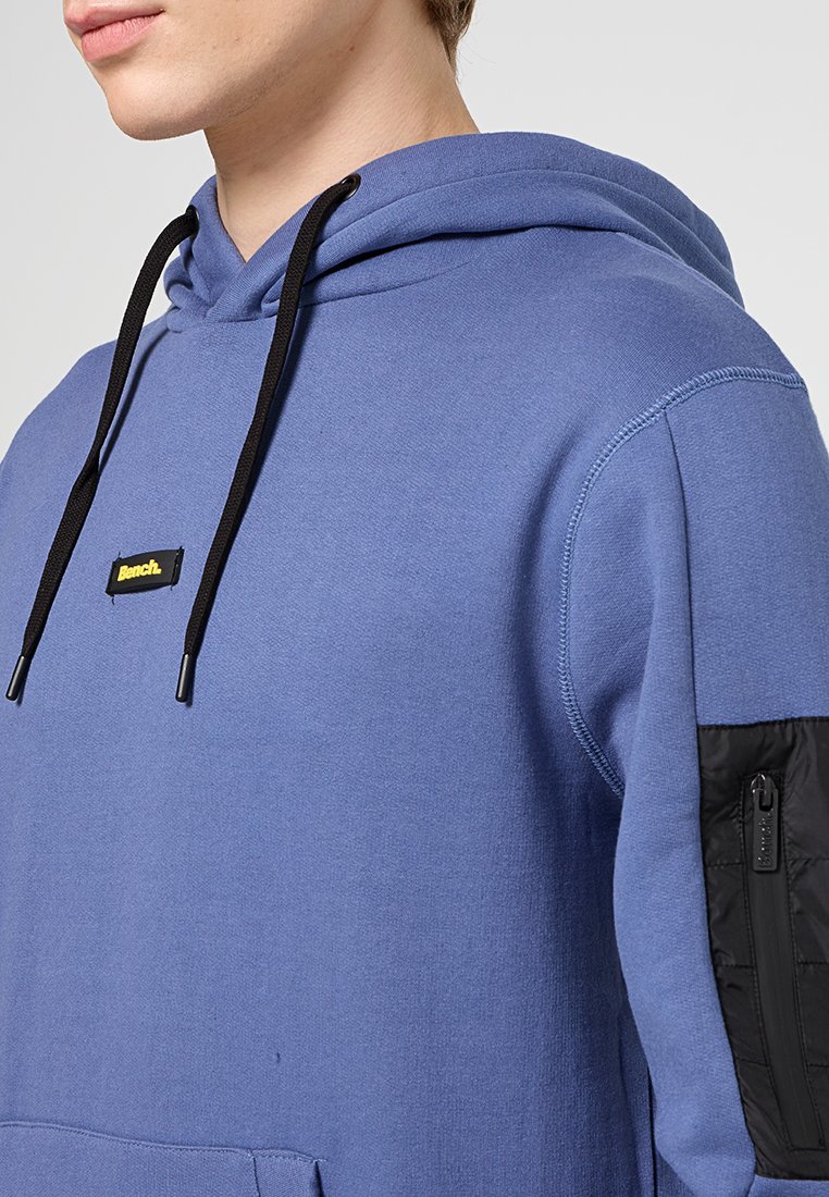 Sudadera azul de mezcla de algodón, cuenta con un bolsillo negro con cremallera en la manga izquierda, cordones ajustables negros y un pequeño parche con el logo.