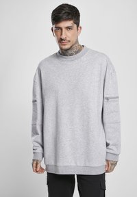 Sudadera gris con cuello redondo, mangas largas y un bolsillo con cremallera en el brazo izquierdo. Textura suave y corte casual.