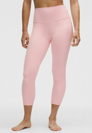 Persona con leggins cortos de talle alto color rosa claro, de pie descalza contra un fondo claro y liso.