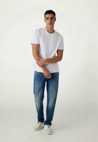 T-shirt branca de gola redonda, jeans de ganga azul e ténis brancos. Modelo está com os braços cruzados, exibindo um visual casual e o tom claro dos jeans.