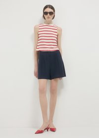 Top senza maniche bianco e rosso a righe, abbinato a pantaloni corti blu navy e scarpe slip-on rosse a punta. Il modello indossa occhiali da sole neri angolari.