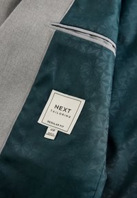 Doublure de costume à motif vert foncé avec une étiquette blanche indiquant « NEXT Tailoring, Regular Fit, 40R, Made in Bangladesh » sur un tissu gris.
