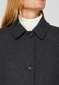 Abrigo de lana gris con cuello tipo solapa, que presenta dos botones y un acabado texturizado. Llevado sobre un cuello de tortuga blanco de canalé.