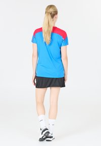 Sportig blå idrottströja med en röd och blå design på axlarna, parat med en svart kjol. Modell bär vita strumpor och svarta skor.