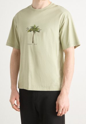 Osoba nosząca jasnozielony T-shirt z małą grafiką palmy i napisem na piersi, w zestawieniu z czarnymi spodniami.