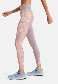 Legginsy z wysokim stanem w jasnoliliowym kolorze, ozdobione wyrazistymi pomarańczowymi wzorami kwiatowymi oraz kontrastowymi bocznymi szwami. Połączone z jasnoniebieskimi butami do biegania.