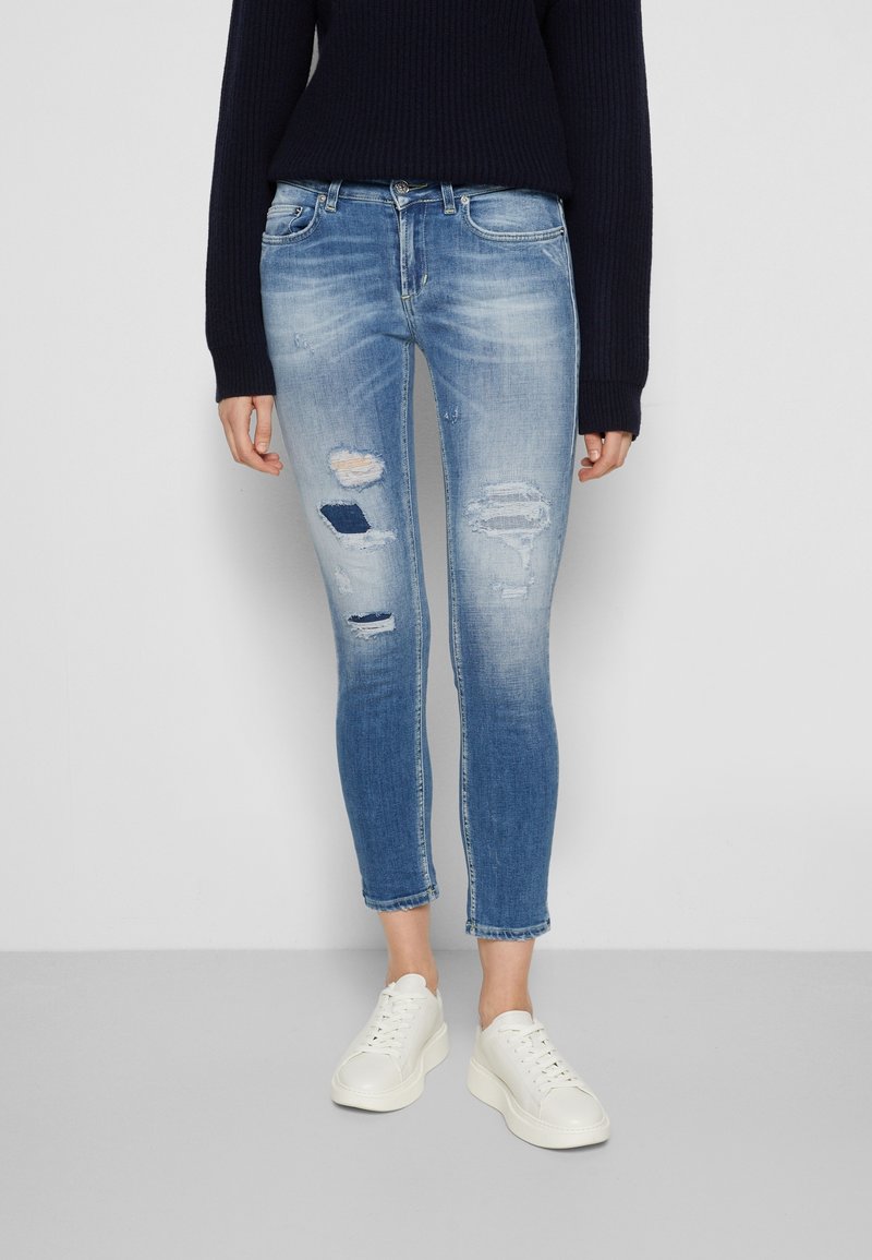 Osoba oblečená v modrých roztrhaných skinny džínách, bílých teniskách a černém svetr s dlouhým rukávem, stojící na pozadí s jednoduchým světlým pozadím.