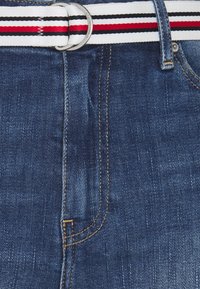 Farkkushortsit, joissa on tummansininen pesu, kultaiset ompeleet ja valkoinen joustovyö, jossa on punaisia ja mustia raitoja sekä hopeinen solki.