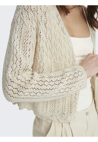 Cárdigan de punto en color blanco roto, con un patrón de tejido abierto, bajo con borde festoneado y mangas anchas. Fabricado con hilo texturizado y ligero.