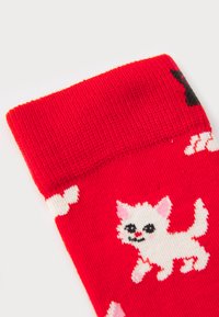 Red knitted socks featuring a white cat design with black accents and pink details, set against a patterned background of clouds.