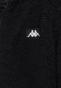 Giacca in pile nera con superficie texturizzata. Caratterizzata da una chiusura a zip e un piccolo logo bianco sulla parte sinistra. Tessuto morbido e accogliente.