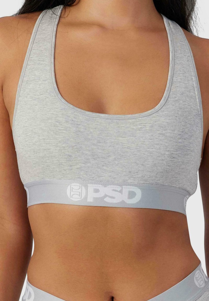 Sujeción deportiva de color gris claro, hecha de una tela suave, que presenta una banda elástica ancha con detalles de logotipo en blanco. Escote en forma curvada y diseño de espalda nadadora.