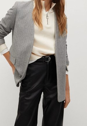 Femme portant un blazer gris, un pull crème à demi-fermeture éclair, une ceinture noire et un pantalon large en cuir noir, les mains détendues le long du corps.