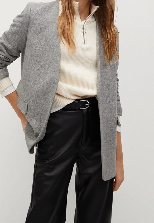 Femme portant un blazer gris, un pull crème à demi-fermeture éclair, une ceinture noire et un pantalon large en cuir noir, les mains détendues le long du corps.