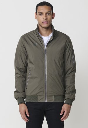 Chaqueta bomber verde oliva con cierre frontal, cintura y puños acanalados. Presenta bolsillos con cremallera en los laterales y en la parte superior del brazo, hecha de un material suave.