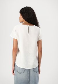 Blusa bianca con maniche corte e tessuto testurizzato. Presenta un'apertura a chiave sul retro e un orlo arrotondato. Abbinata a una gonna di jeans.