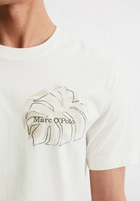 Short-sleeve, cream-colored cotton t-shirt featuring a brown leaf graphic and the text "Marc O'Polo" in a modern font.