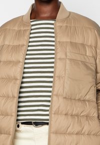 Béžová prošívaná bunda se zipem vpředu, žebrovaným límcem a boční kapsou. Nošena přes pruhované tričko se zelenými a bílými vodorovnými pruhy.