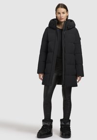 Svart uppblåsbar jacka med huva, med ett quiltat mönster, en frontdragkedja och tryckknappar. Kombineras med svarta leggings och stövlar.