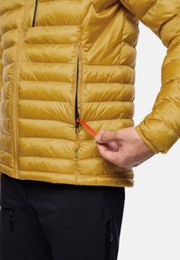 Gelbe Puffjacke mit horizontalen Steppnähten, seitlicher Reißverschlusstasche und einem roten Ziehband mit der Aufschrift "PACK AWAY". Schwarze Hose im Hintergrund.