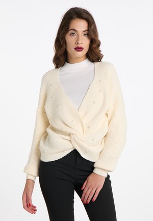 Vrouw met golvend bruin haar die een witte coltrui en een gedraaide crèmekleurige trui met kleine parels draagt, gecombineerd met een zwarte broek.