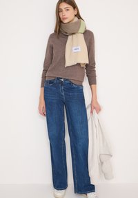 Blå viddebukser kombineret med en brun sweater og et flerfarvet tørklæde. Tørklædet har en beige bund med grønne og brune striber.