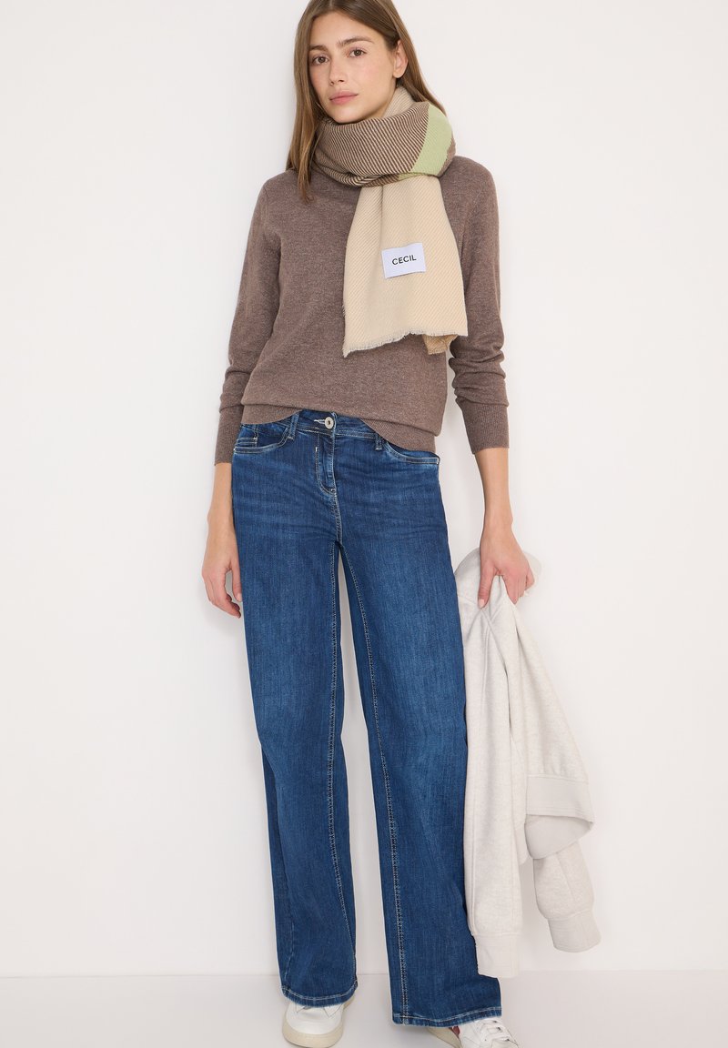 Blå viddebukser kombineret med en brun sweater og et flerfarvet tørklæde. Tørklædet har en beige bund med grønne og brune striber.